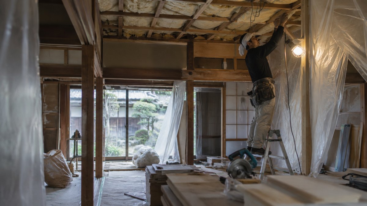 内装をはがしている古い日本家屋の室内。天井板が外されて断熱材と梁が見えており、養生シートが下がっている