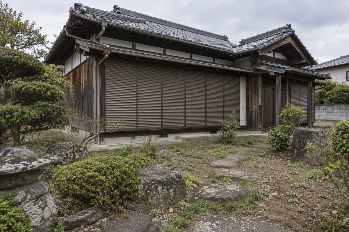 雨戸を閉め切った瓦屋根の木造住宅と、雑草が伸び始めた庭。曇りの日