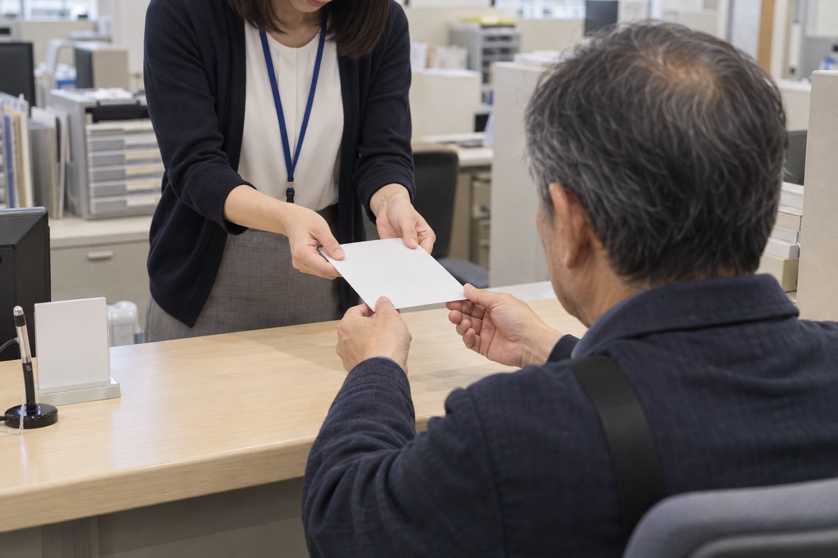 市役所の戸籍の窓口で、カウンター越しに職員から白い封筒を受け取ろうとしている来庁者の後ろ姿