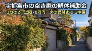 生け垣の伸びた古い木造二階建ての空き家と、その前の細い坂道。奥に住宅が続く