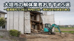 白い養生シートの足場に囲まれた木造二階建ての家を、小型の重機で解体している現場。奥に田畑と低い家並み