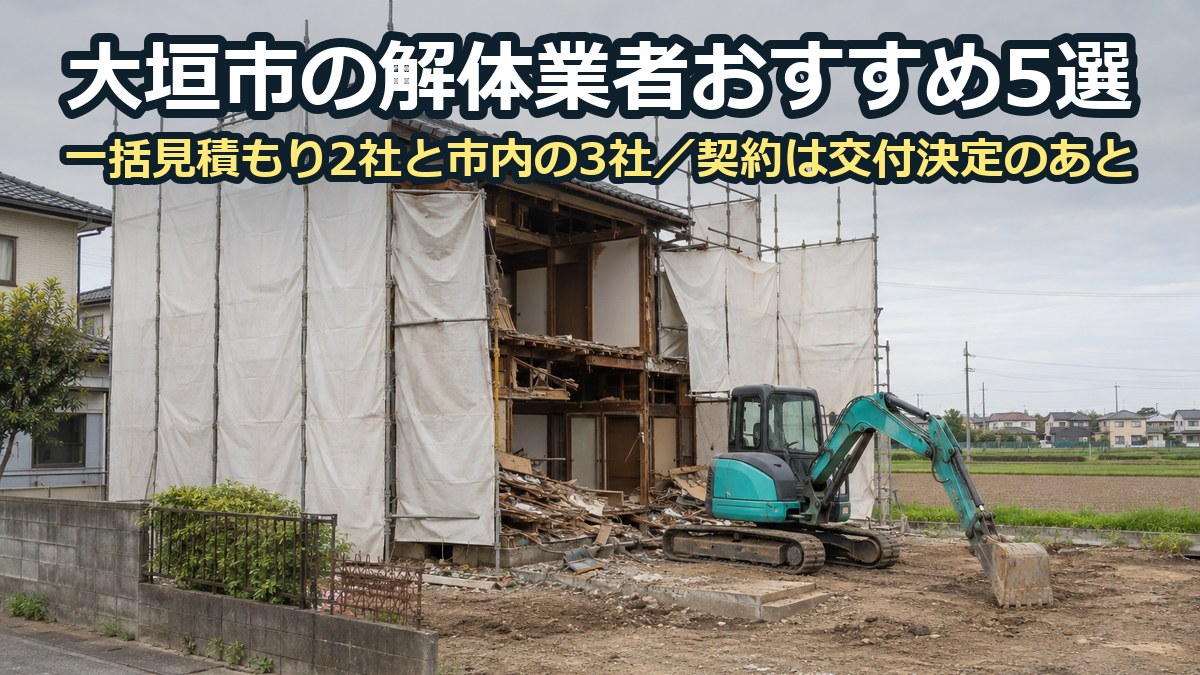 白い養生シートの足場に囲まれた木造二階建ての家を、小型の重機で解体している現場。奥に田畑と低い家並み